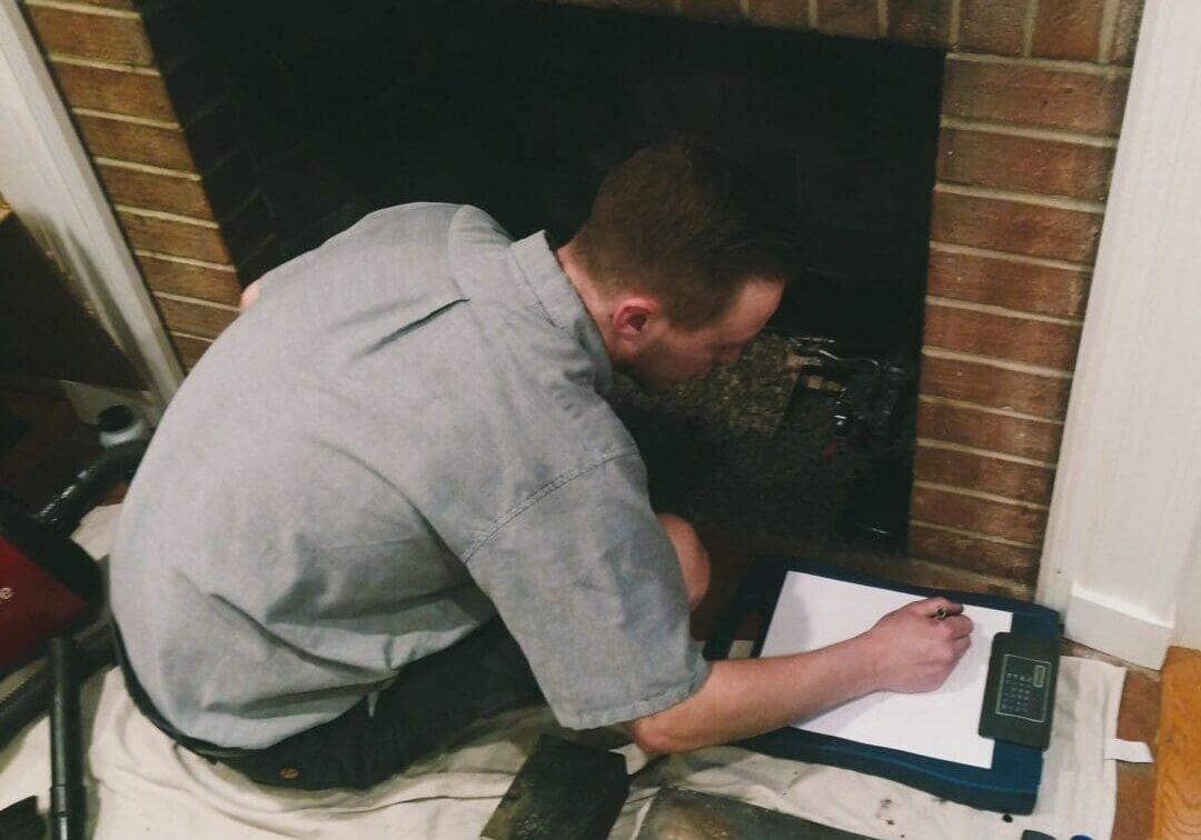 Technician sits in front of a fireplace opening as he inspects and notes the fireplace